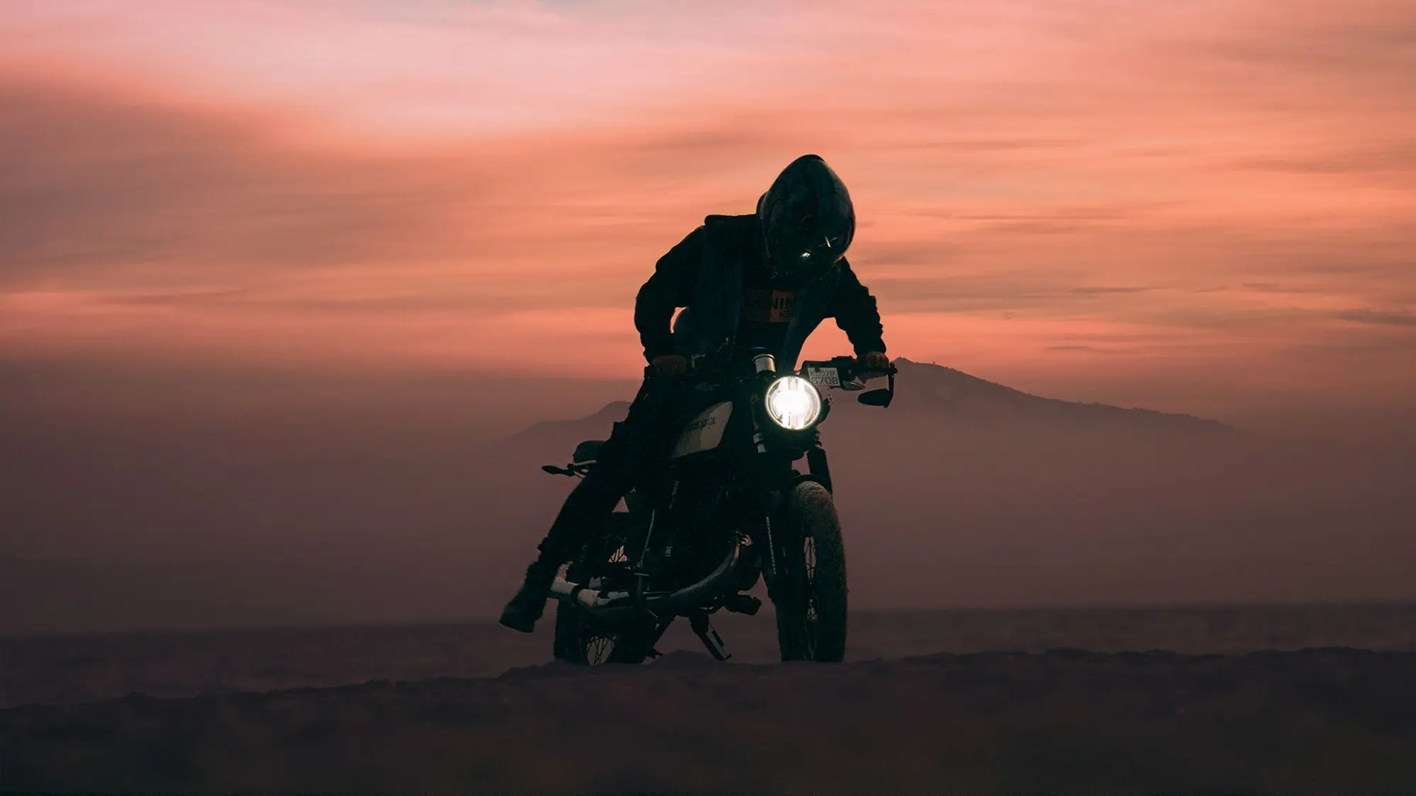 A rider pauses on a motorbike at dusk, headlight on, against a fading sky