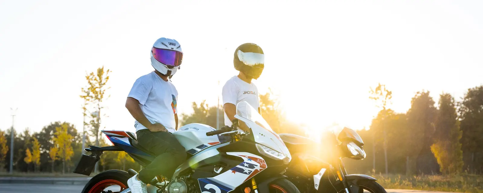 Two riders in helmets stand with their bikes in low evening sun