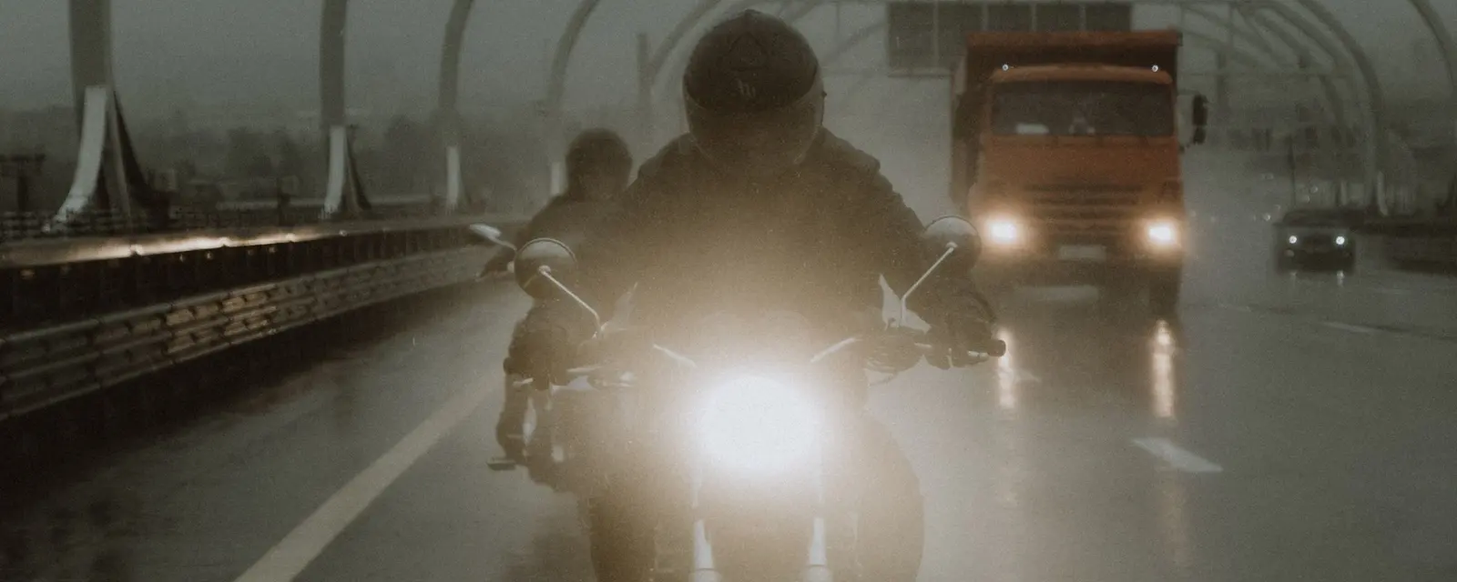 A motorbike headlight cuts through spray on a wet motorway, a lorry behind